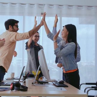 Na sliki skupina sodelavcev v pisarni proslavlja uspeh z dvigom rok in ploskanjem.