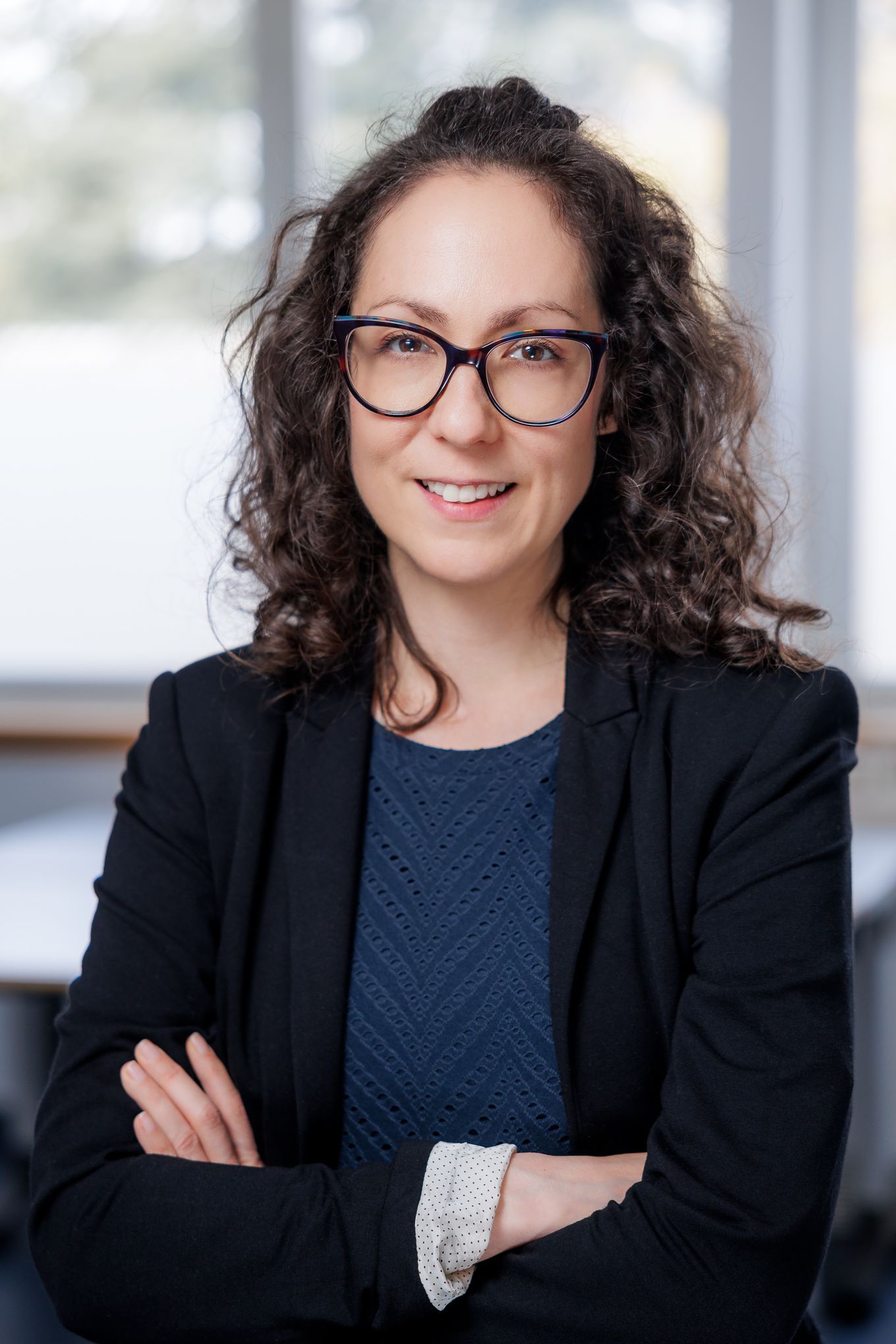 Portret nasmejane ženske z očali, sklenjenimi rokami in temnim suknjičem v pisarni.