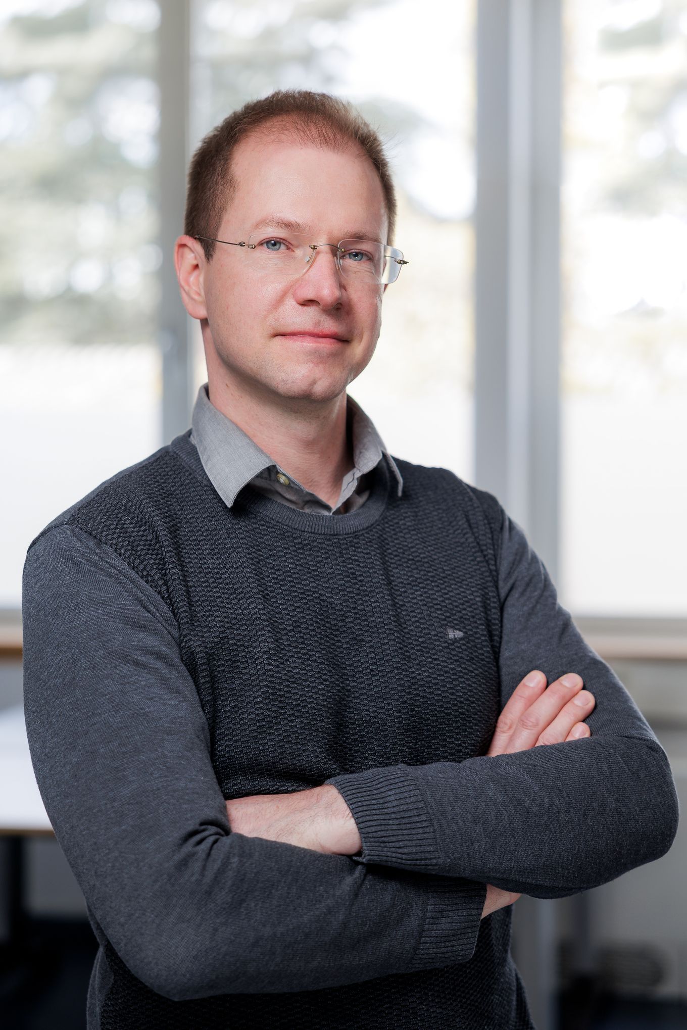 Portret moškega z očali in prekrižanimi rokami v sivem puloverju v pisarniškem prostoru.