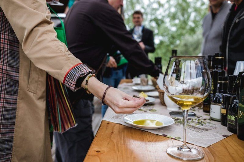 Fotografija mize, na kateri so razstavljena jedilna olja različnih tipov in kozarec vina. Poleg mize je skupina obiskovalcev, kateri poskušajo različne dobrote.