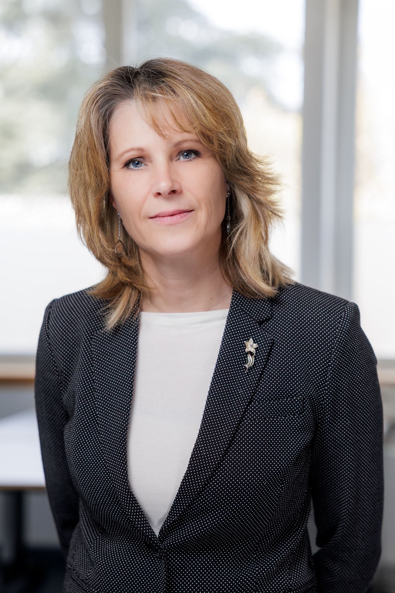 Portret ženske v temnem suknjiču in beli majici v svetlem pisarniškem prostoru.